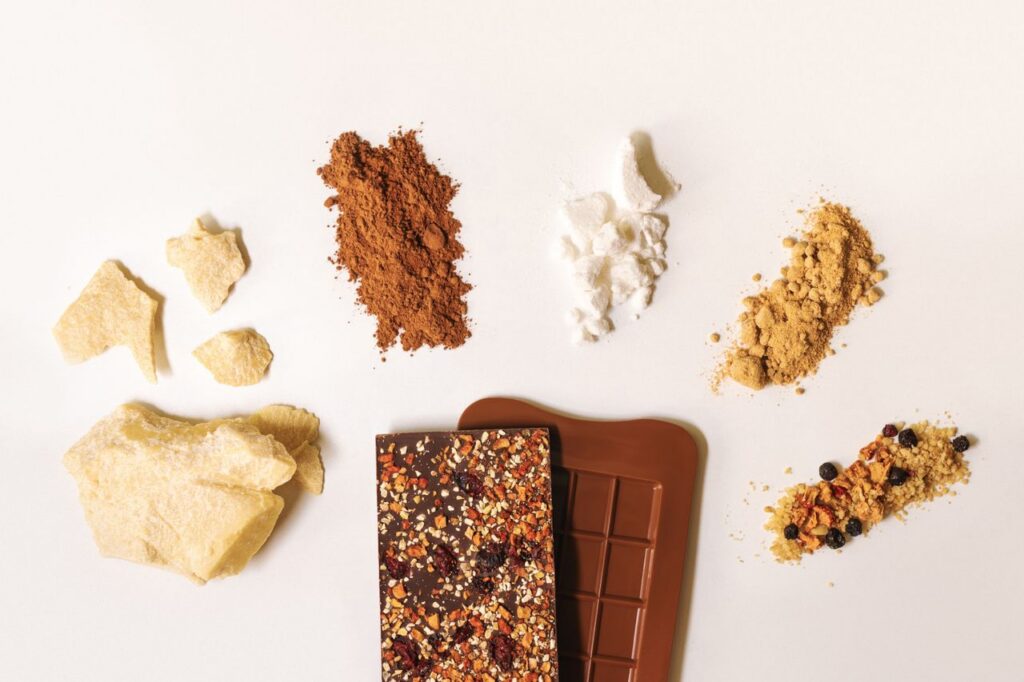 Flat lay of artisan chocolate bar surrounded by raw ingredients on a white background.